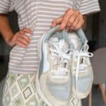 Person in a striped shirt and patterned skirt holding a pair of worn sneakers by the laces indoors.