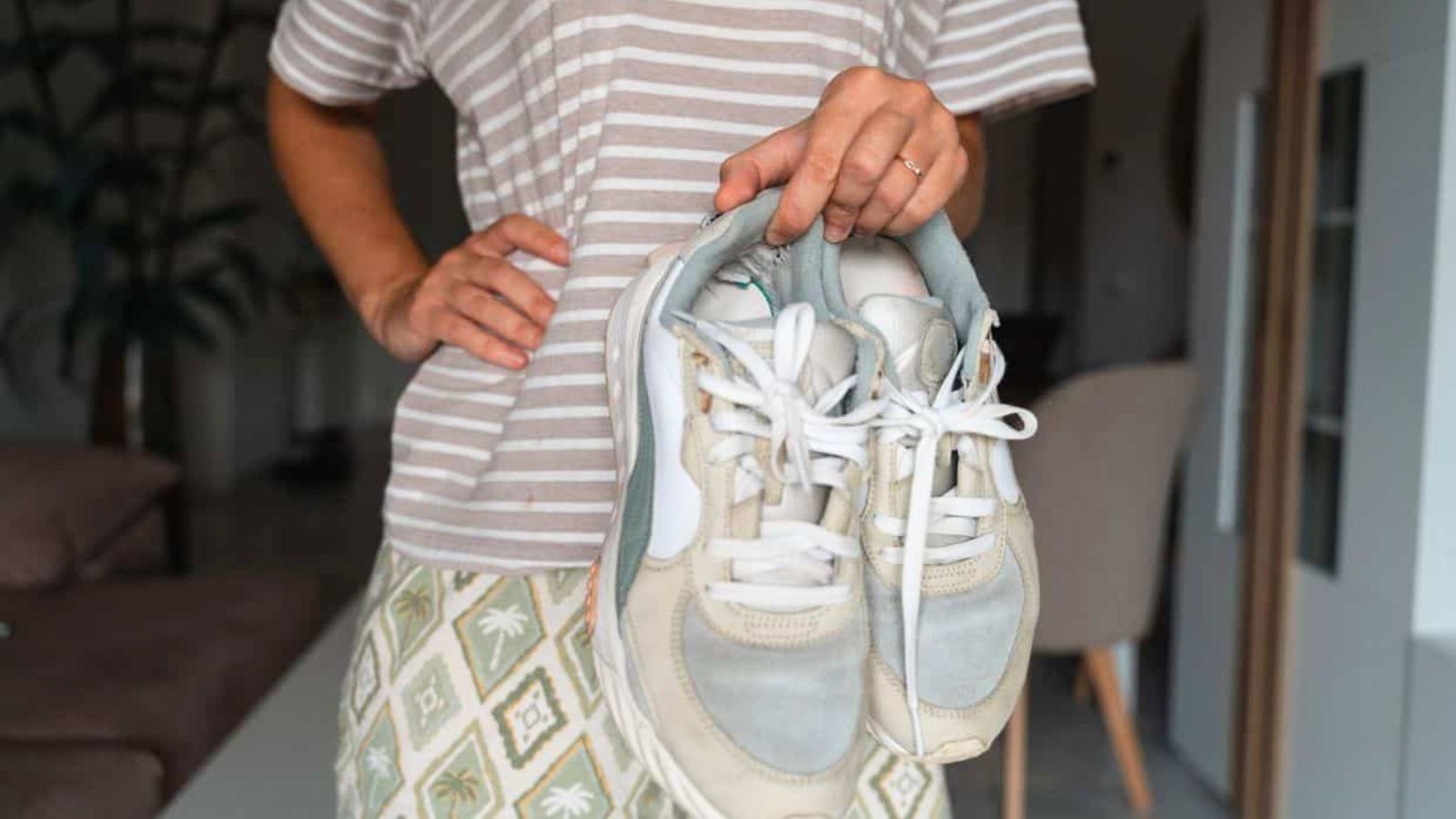 Person in a striped shirt and patterned skirt holding a pair of worn sneakers by the laces indoors.