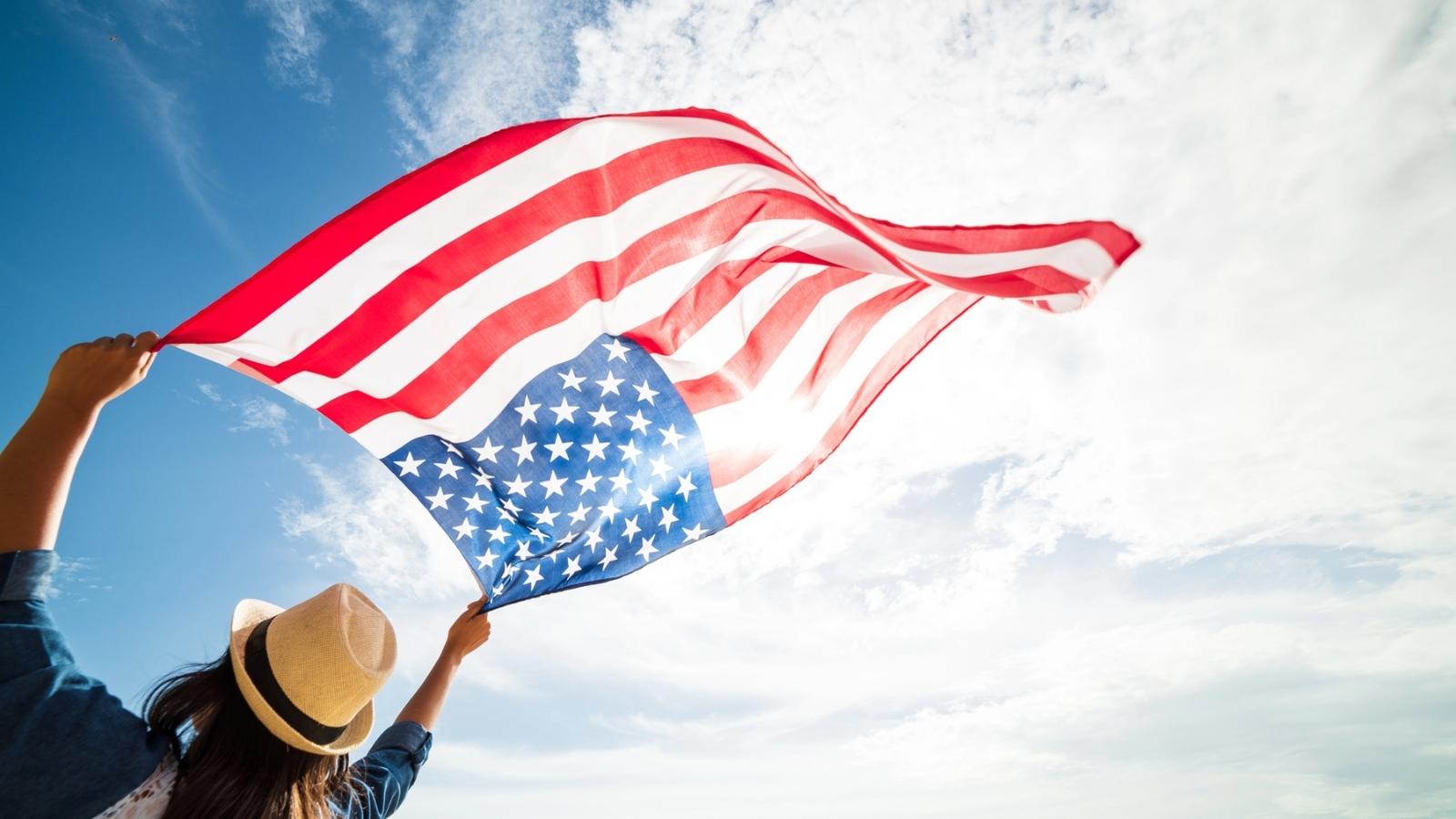 A person wearing a hat holds up a large American flag outdoors against a blue sky with clouds, celebrating the spirit of American food cities.