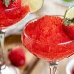 A glass filled with red shaved ice, garnished with a lime slice and a whole strawberry, with more strawberries and lime in the background—perfect for celebrating National Daiquiri Day.