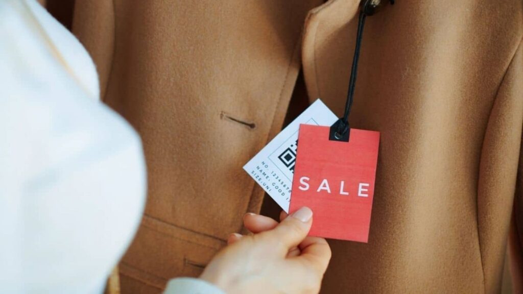 A hand holds a coat&rsquo;s sale tag and a QR code tag attached to a brown coat, reflecting the mindful choices of simple living.
