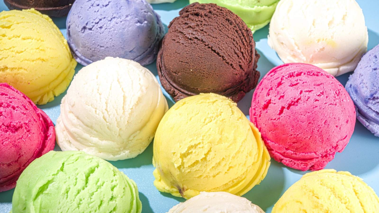 Scoops of colorful ice cream, including pink, yellow, green, purple, white, and brown, are artfully arranged on a light blue surface—perfect for celebrating National Ice Cream Day.