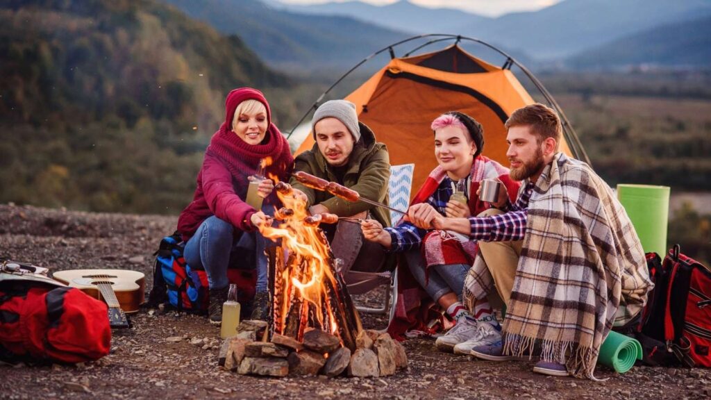 Four people sit by a campfire in front of an orange tent, enjoying the simplicity of analog camping as they roast food on sticks. Camping gear and mountains are visible in the background.