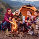 Four people sit by a campfire in front of an orange tent, enjoying the simplicity of analog camping as they roast food on sticks. Camping gear and mountains are visible in the background.