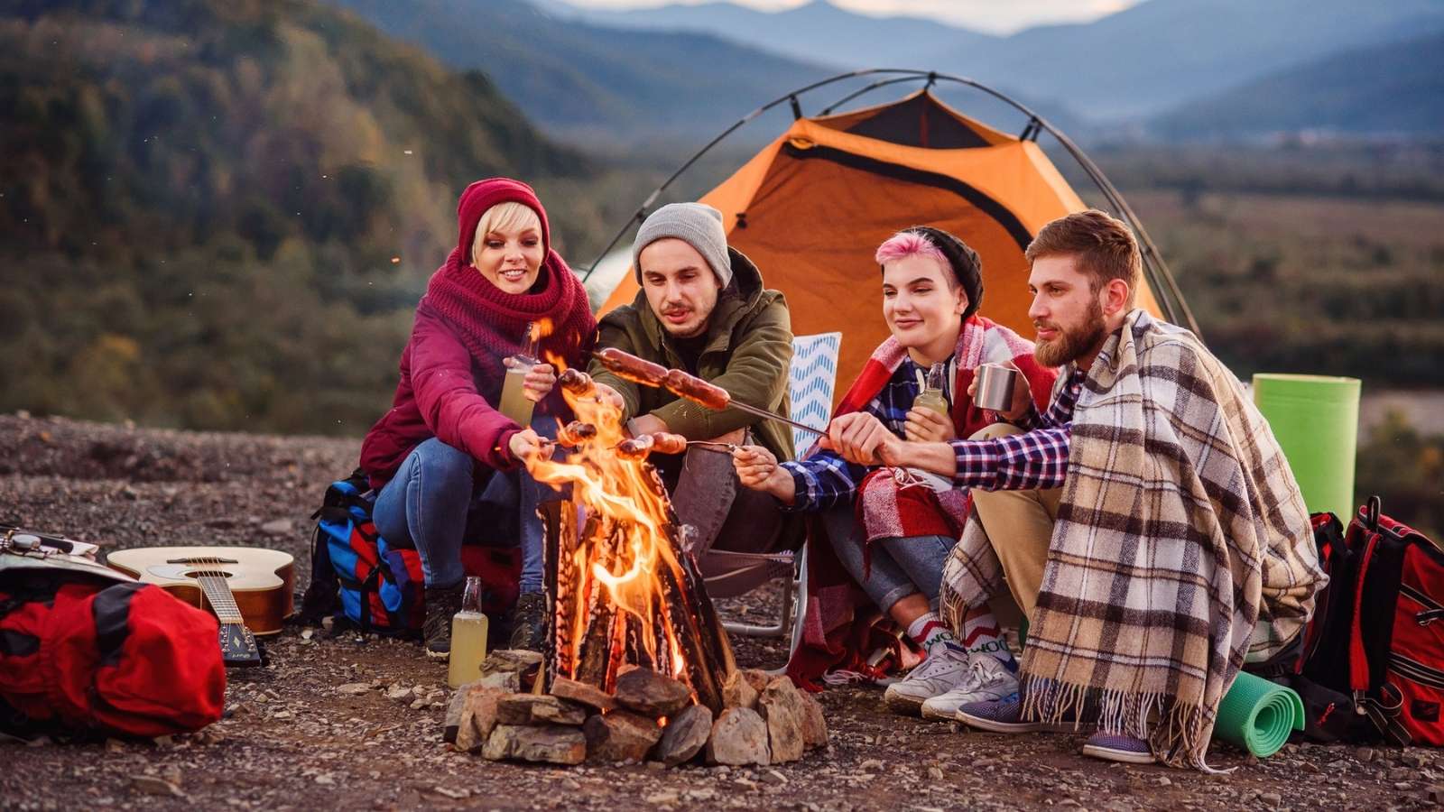 Four people sit by a campfire in front of an orange tent, enjoying the simplicity of analog camping as they roast food on sticks. Camping gear and mountains are visible in the background.