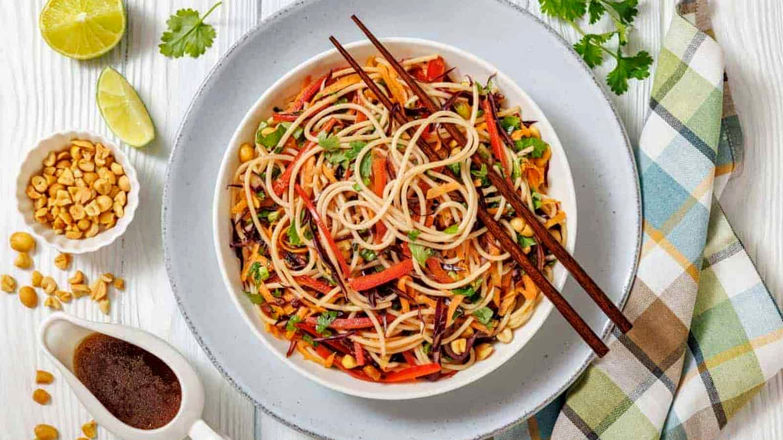 A bowl of Asian noodle salad with vegetables and chopsticks, surrounded by lime, peanuts, sauce, and fresh cilantro on a light wooden table with a plaid napkin.