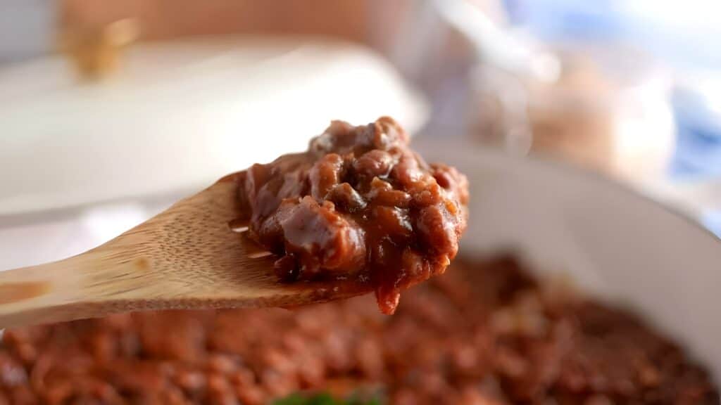 A wooden spoon holds a serving of thick, chunky chili above a pot filled with more of the same dish.