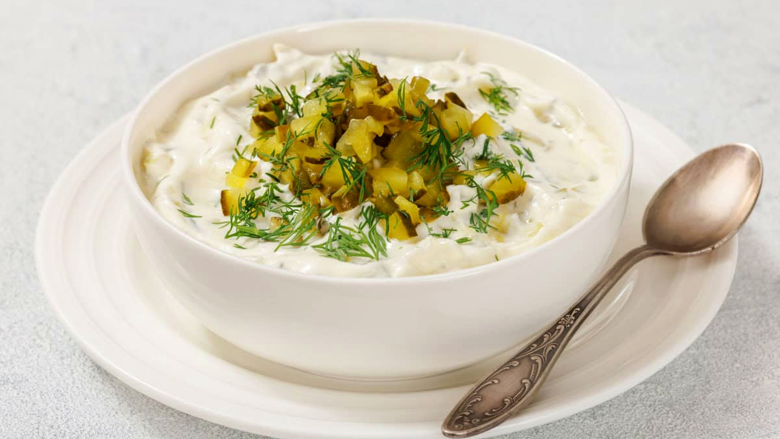 A bowl of Dill Pickle Dip with chopped pickles and dill on top, placed on a white plate. A spoon rests beside the bowl.