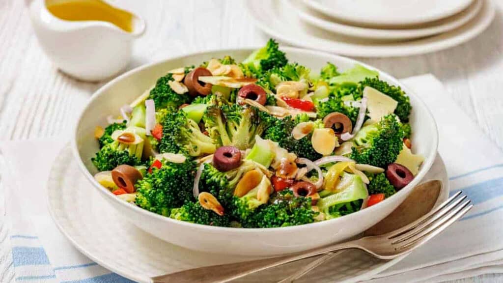 A bowl of broccoli salad with olives, sliced almonds, onions, and red peppers, served with a fork and spoon on a white table.