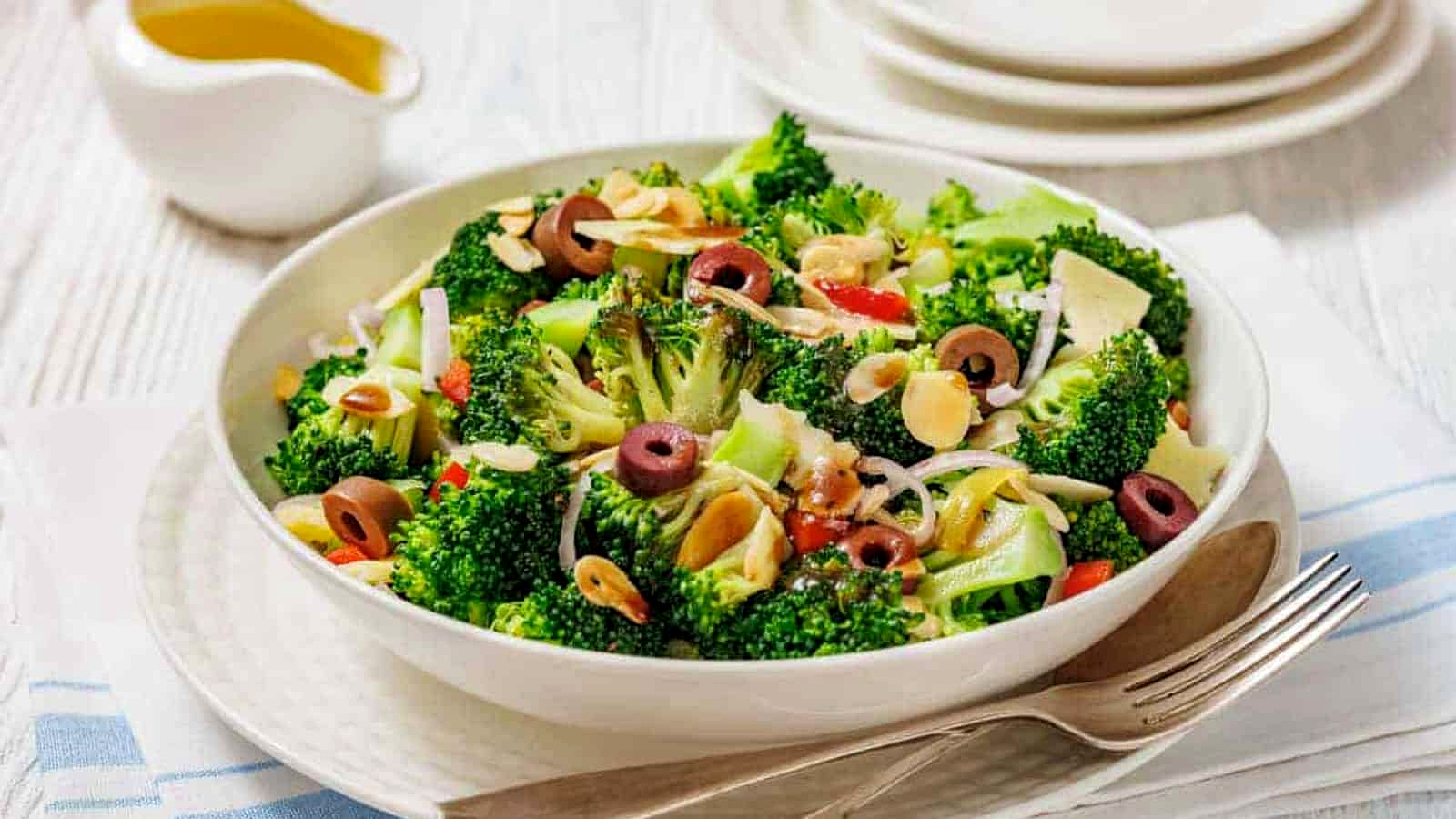 A bowl of Italian broccoli salad with sliced olives, almonds, onions, and red peppers, served on a white plate with a fork and napkin beside it.