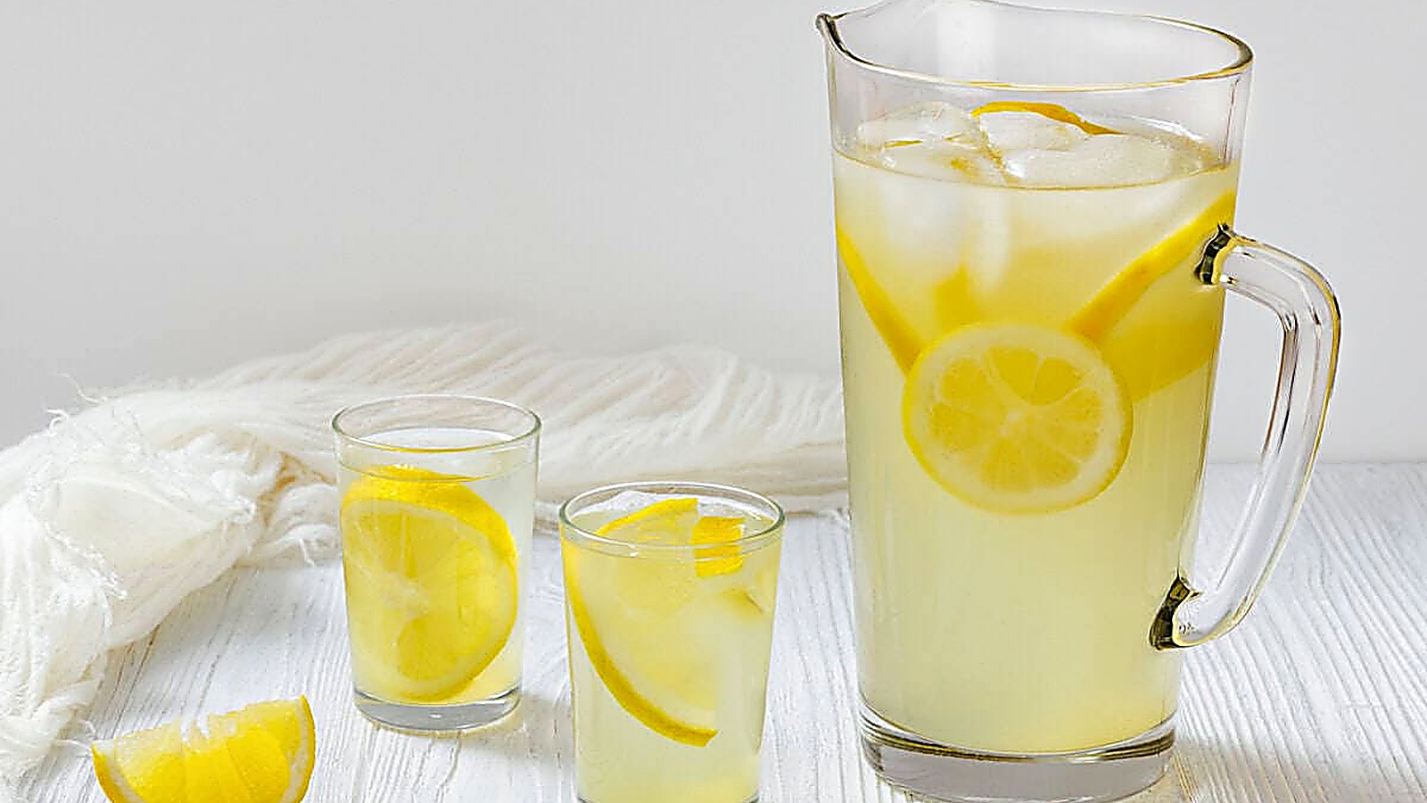 A tall glass pitcher filled with lemonade, ice, and lemon slices stands on a white wooden table, accompanied by two smaller glasses also containing lemonade and lemon. A lemon wedge is placed beside the glasses. A white cloth is draped in the background.