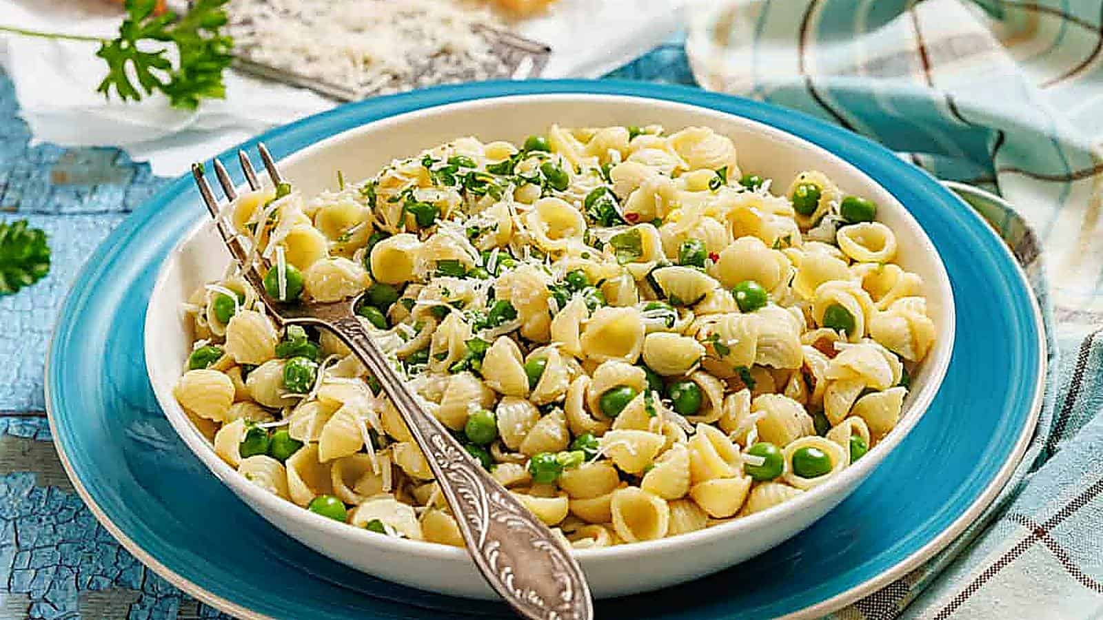 A bowl of pasta and peas with a fork, grater, and cheese on the side.