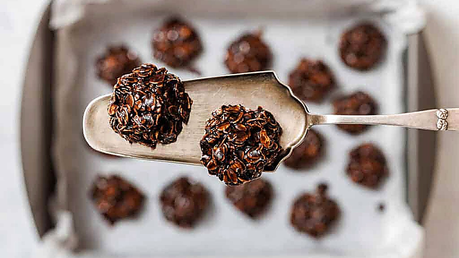A metal spatula holds two chocolate Peanut Butter No-Bake Cookies above a baking tray lined with parchment paper and more cookies.