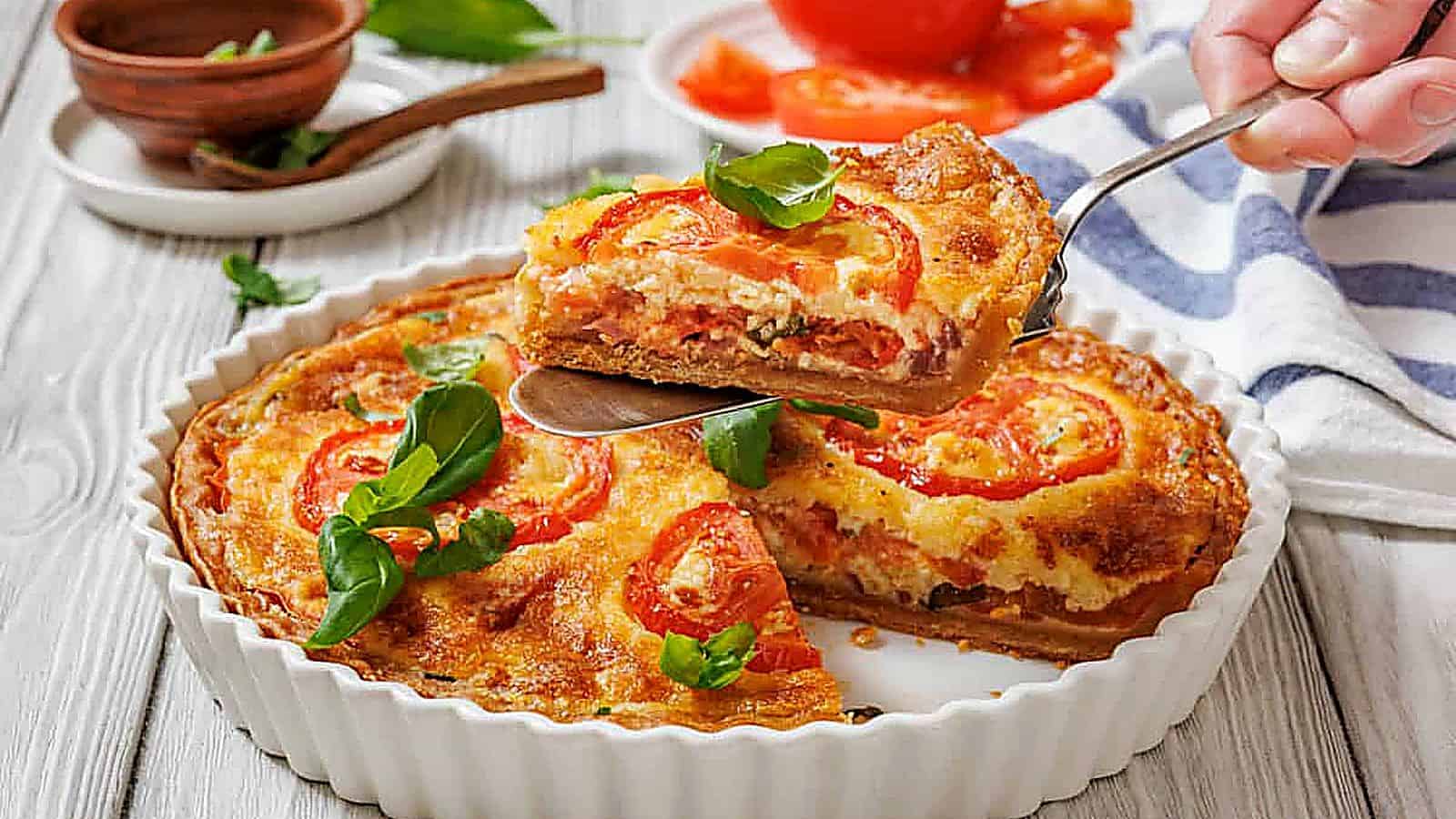 A slice of tomato pie being lifted with a serving utensil from a white fluted dish. A whole tomato pie is visible in the dish, garnished with tomato slices and basil.