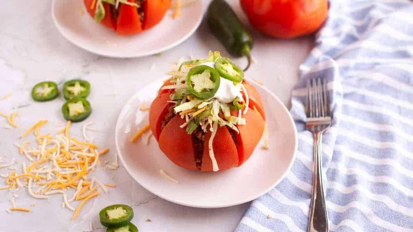Two taco stuffed tomatoes on white plates with toppings and a fork.