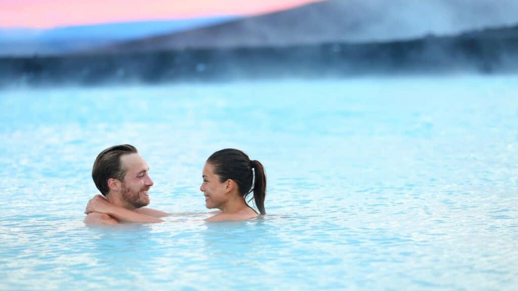A couple stands close together in a large outdoor hot spring, facing each other and smiling, with steam rising in the background.