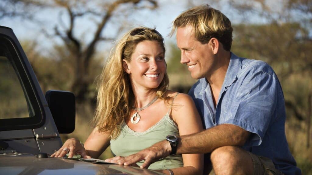 A woman and man sit outdoors next to a vehicle, smiling at each other during their safari honeymoon. Trees and dry grass are visible in the background.