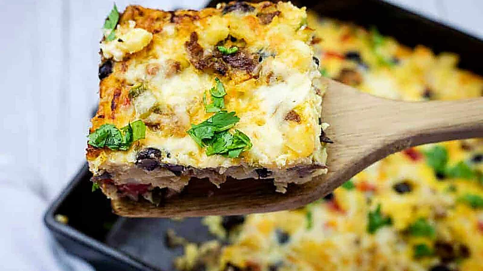A wooden spatula holds up a square slice of baked casserole topped with melted cheese and chopped herbs, with more casserole in the background.