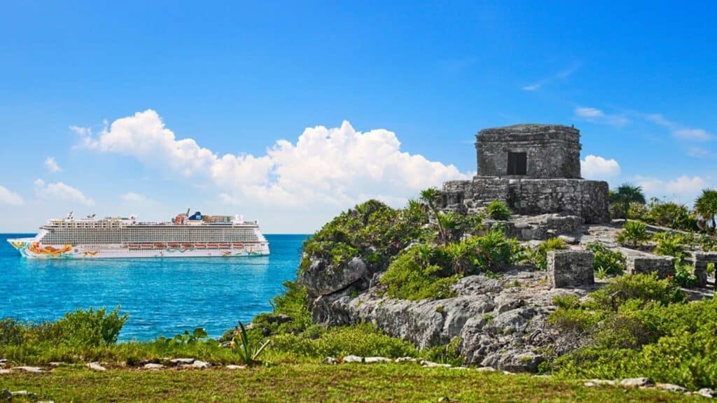 A large white cruise ship, possibly part of Sixthman Cruises, sails near the coastline, with an ancient stone ruin and greenery in the foreground under a bright blue sky.