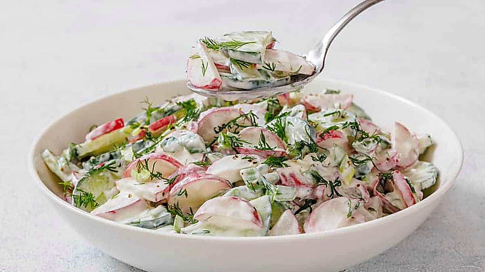 A bowl of Creamy Radish Salad garnished with dill, with a spoon lifting a portion above the bowl.