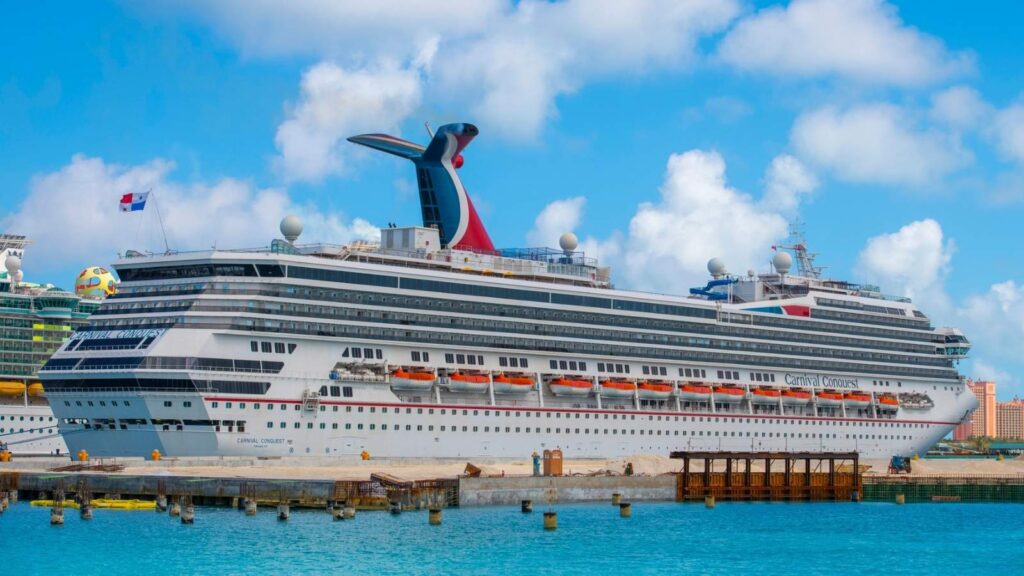 A large white cruise ship is docked at a port in clear blue water under a partly cloudy sky. The ship has multiple decks and red, white, and blue funnel.