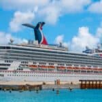 A large white cruise ship is docked at a port in clear blue water under a partly cloudy sky. The ship has multiple decks and red, white, and blue funnel.