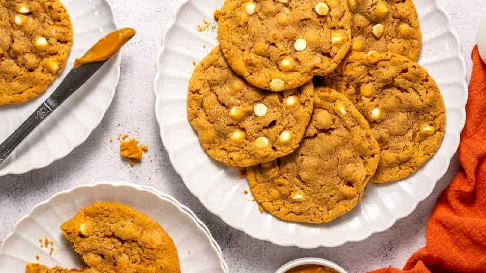 A plate of large peanut Copycat Crumbl Biscoff cookies with white chocolate chips, surrounded by a knife, a small bowl of peanut butter, and an orange napkin.