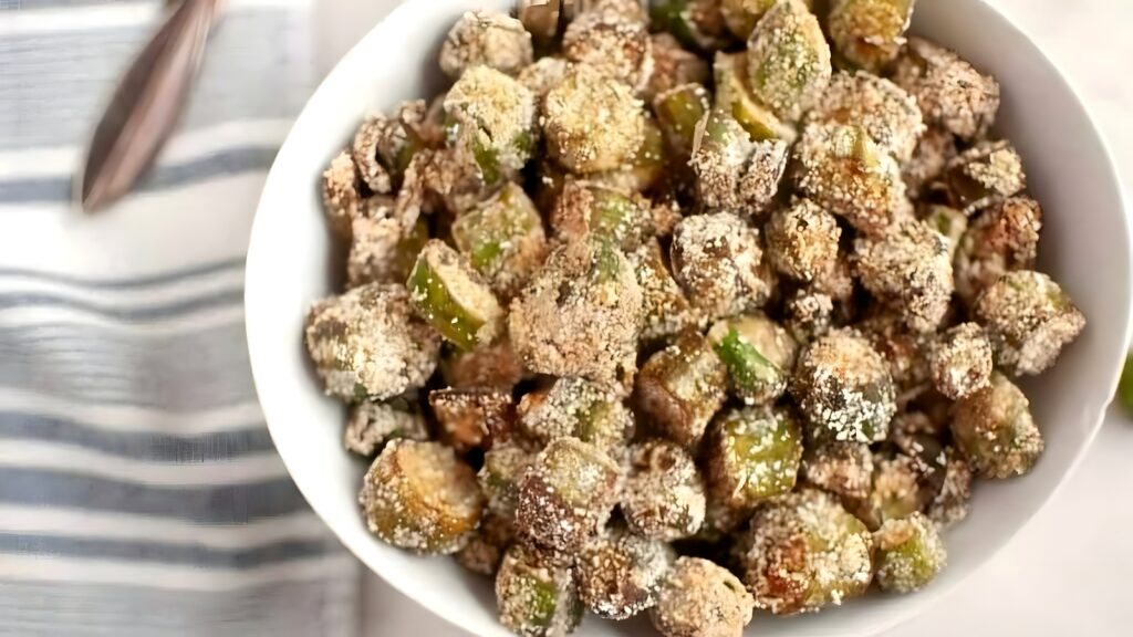 A white bowl filled with breaded, seasoned fried okra pieces sits on a white surface next to a striped cloth napkin and a fork.