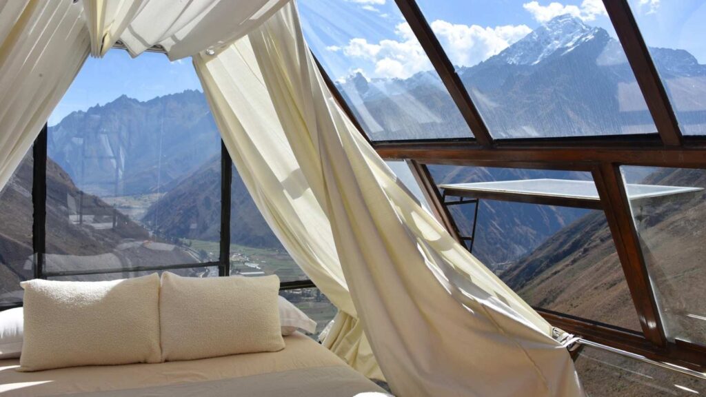 A bed with cream pillows and sheets is positioned next to large glass windows, offering a view of mountains and a valley under a partly cloudy sky&mdash;perfect for those seeking unusual stays.