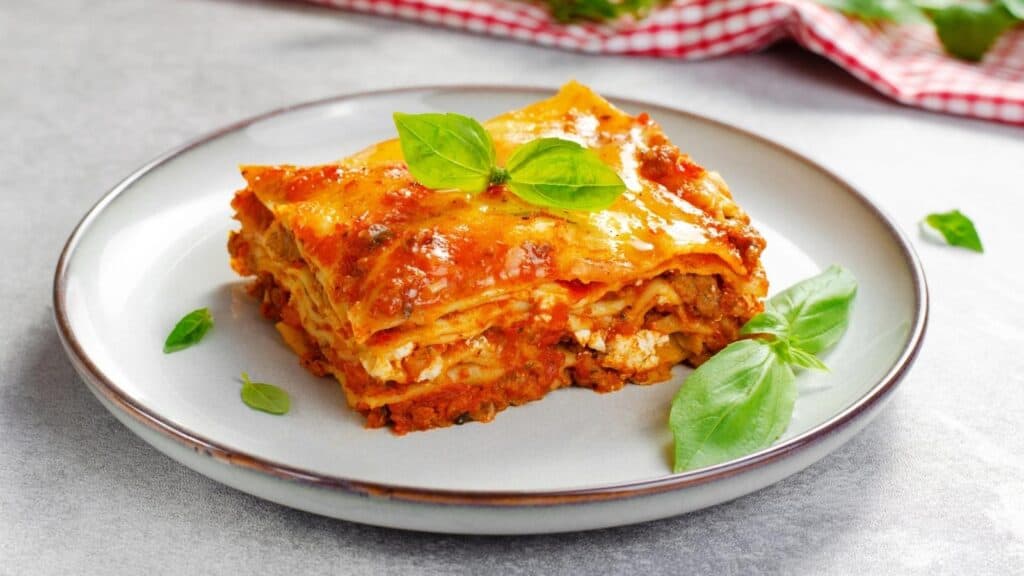 A slice of lasagna with tomato sauce and cheese sits on a white plate, garnished with fresh basil leaves&mdash;an irresistible example of classic lasagna recipes.