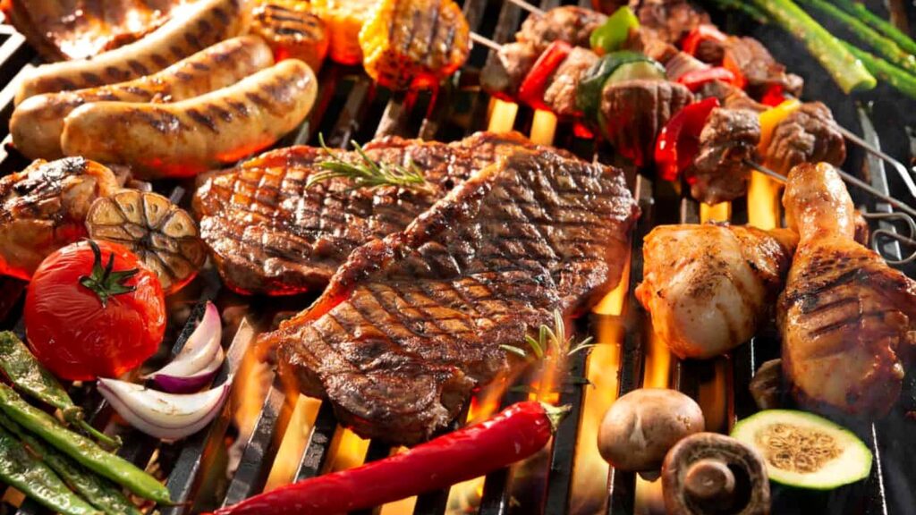 Various meats and vegetables, including steak, sausages, kebabs, mushrooms, and peppers, cooking on a grill with visible flames&mdash;a mouthwatering scene perfect for anyone seeking grilling tips for beginners.
