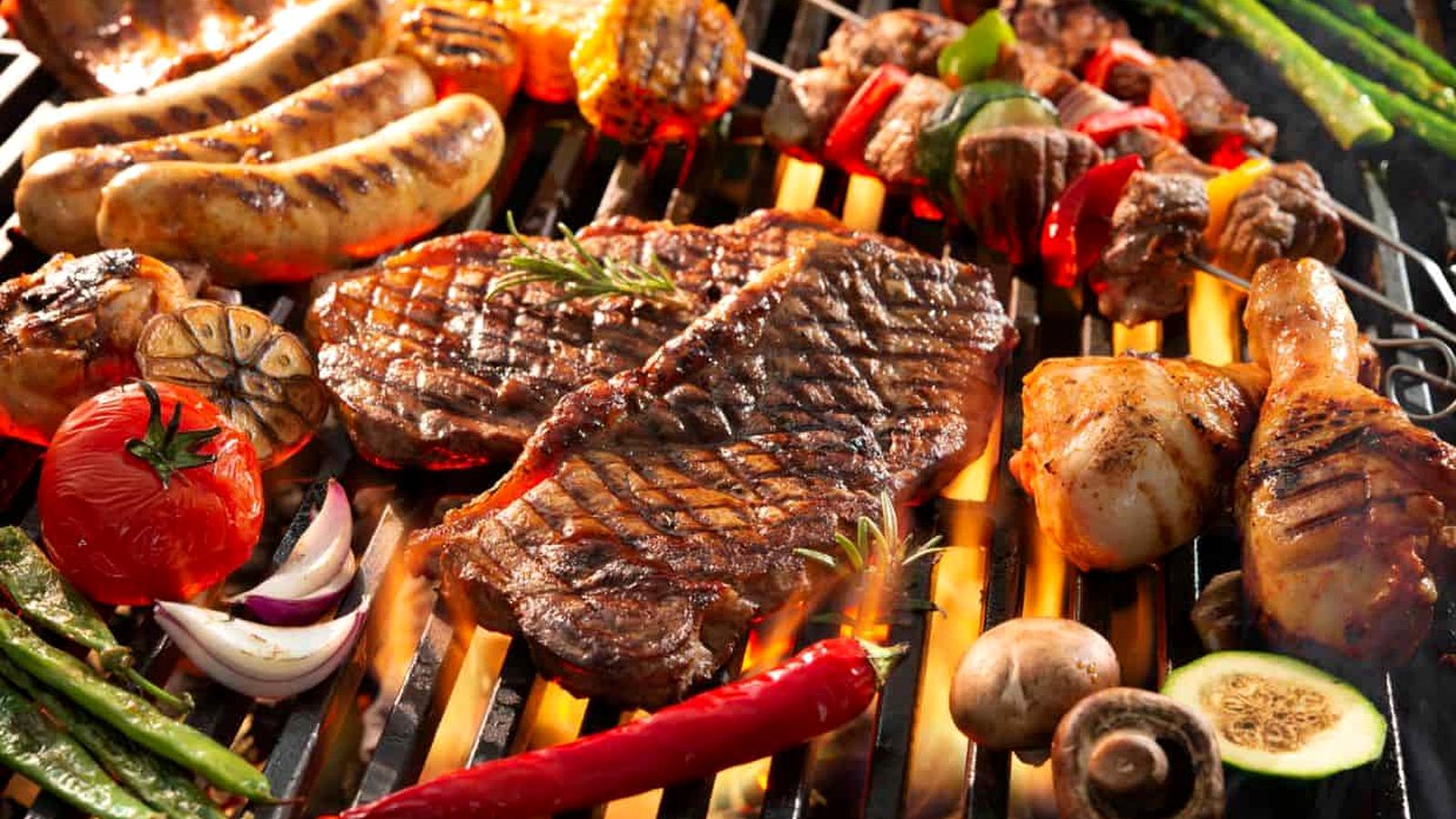 Various meats and vegetables, including steak, sausages, kebabs, mushrooms, and peppers, cooking on a grill with visible flames—a mouthwatering scene perfect for anyone seeking grilling tips for beginners.
