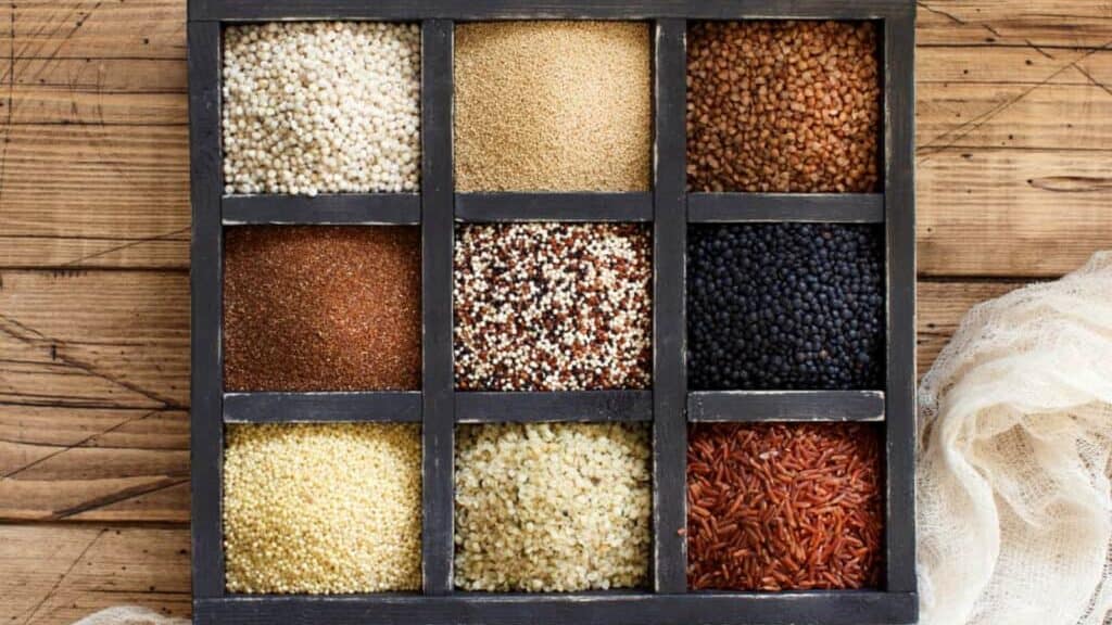 A wooden box with nine sections, each filled with a different type of grain or seed, arranged neatly on a wooden surface.