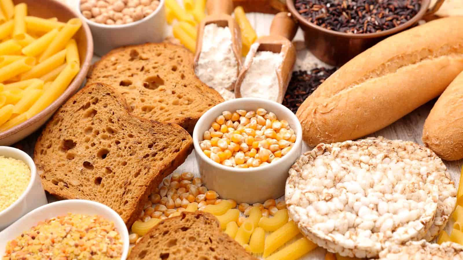 Assorted carbohydrate-rich foods including pasta, bread, rice cakes, popcorn kernels, flour, beans, and sliced toast displayed on a light surface.