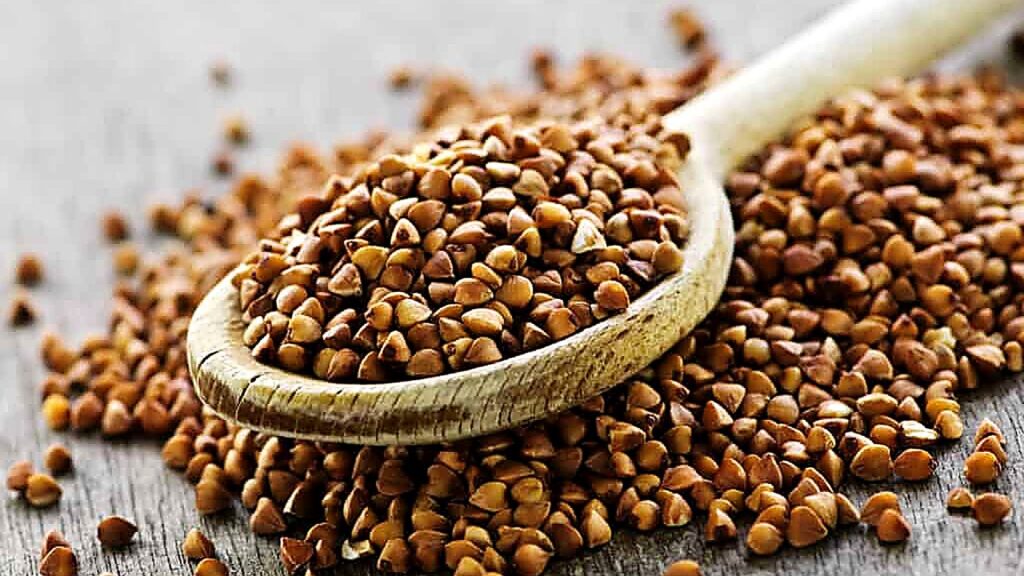 A wooden spoon filled with raw buckwheat groats rests on a wooden surface, with more groats scattered around it.