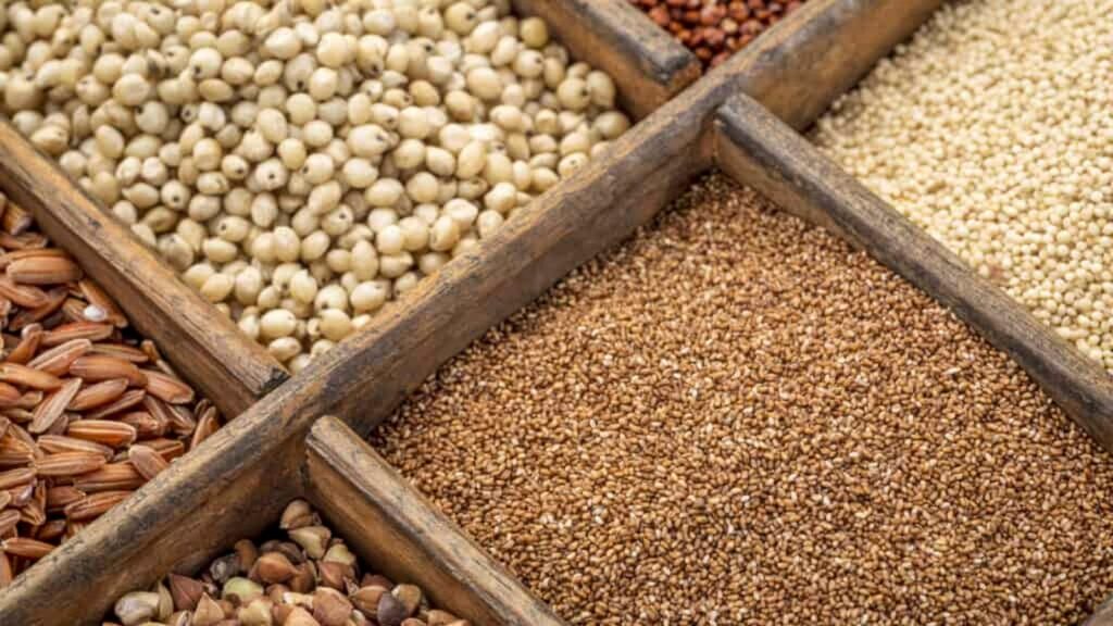 Six sections of a wooden tray display assorted grains, including sorghum, millet, brown rice, and seeds, viewed from above.