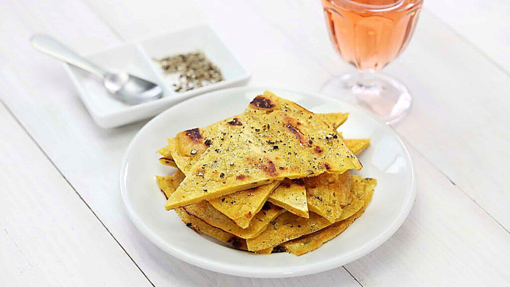 A white plate with crisp flatbread pieces, sprinkled with black pepper, next to a glass of pink drink and a dish with salt and pepper on a white table.