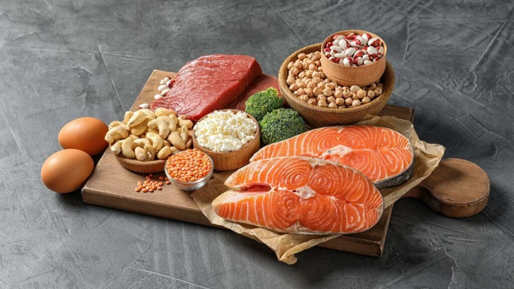 Assorted protein-rich foods, including raw salmon, beef, eggs, chickpeas, nuts, cottage cheese, lentils, and broccoli, arranged on a wooden board on a gray surface.