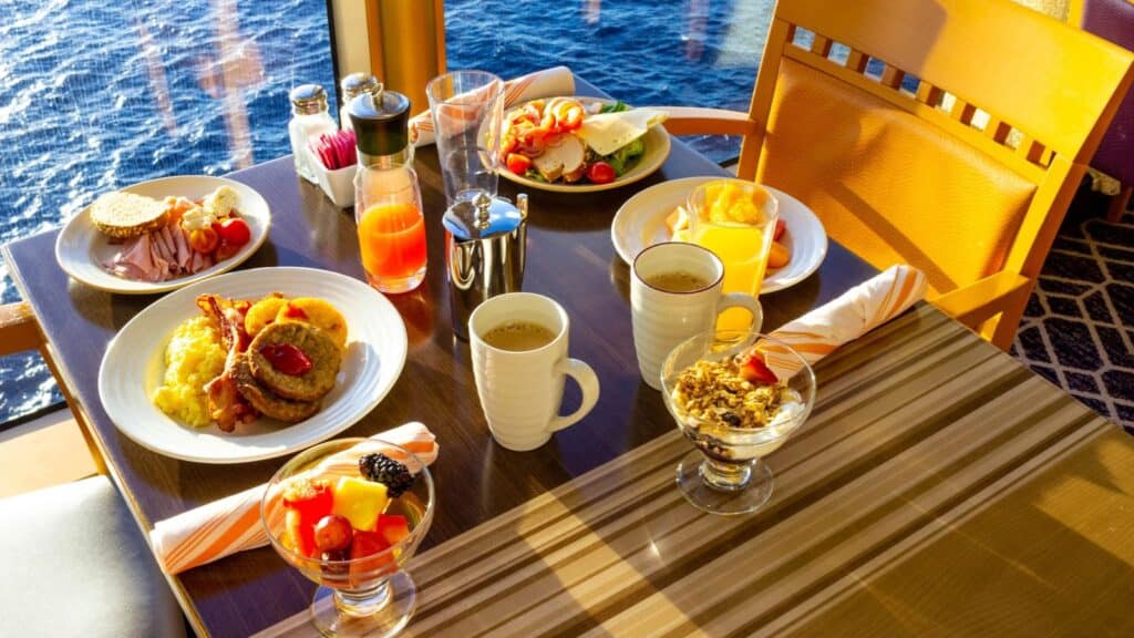 Breakfast spread on a table by a window with ocean view, featuring eggs, pancakes, fruit, juice, coffee, cereal, and condiments.