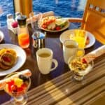 Breakfast spread on a table by a window with ocean view, featuring eggs, pancakes, fruit, juice, coffee, cereal, and condiments.