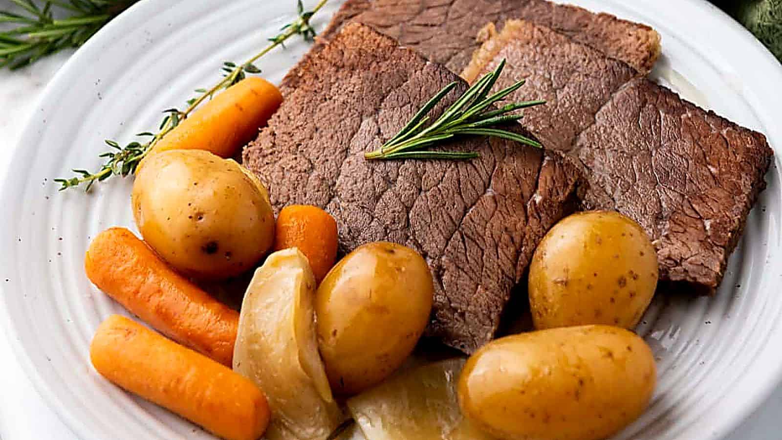 A plate of slow cooker rump roast with potatoes, carrots, onions, and a sprig of rosemary on a white dish.