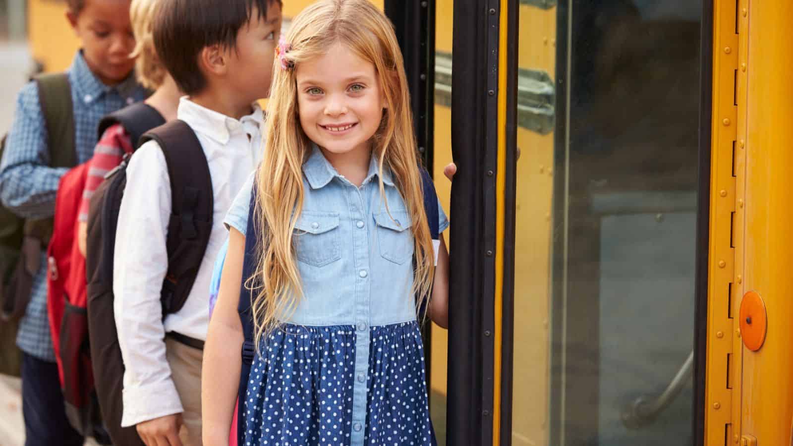 A young girl with long blond hair smiles while boarding a yellow school bus, with other children waiting in line behind her.