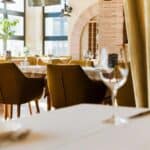 A modern restaurant dining area with green chairs, set tables, wine glasses, large windows, and natural light streaming in.