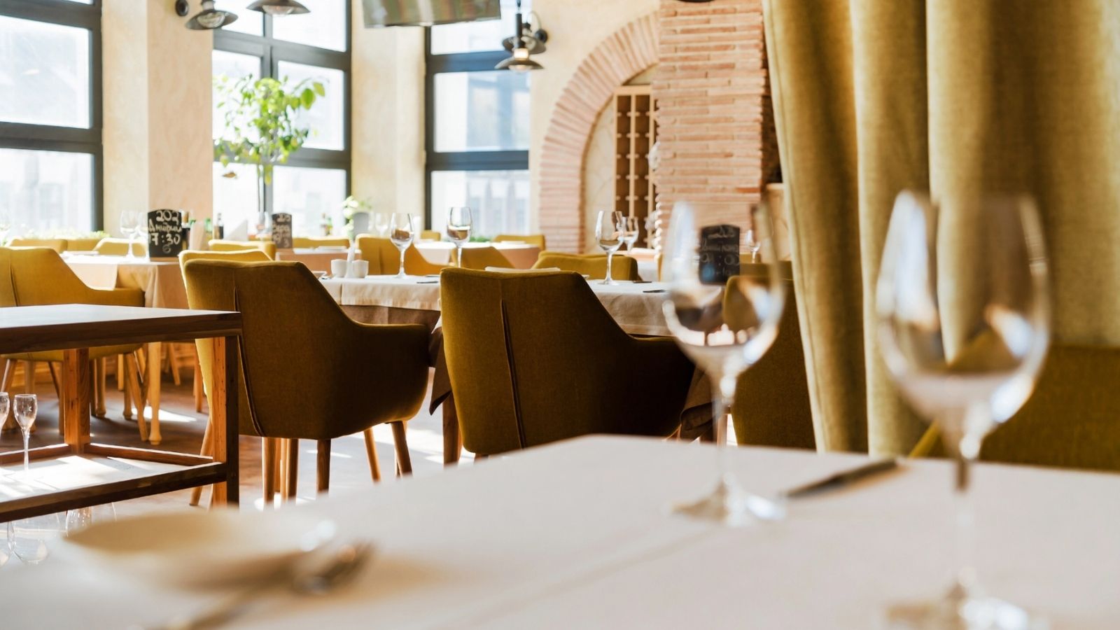 A modern restaurant dining area with green chairs, set tables, wine glasses, large windows, and natural light streaming in.