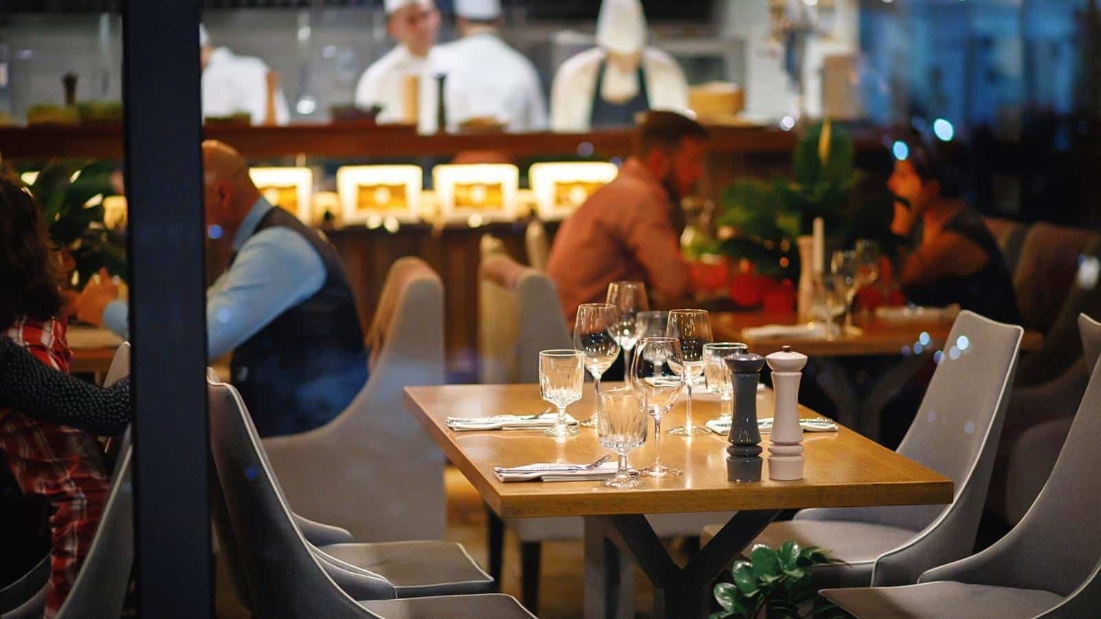 A restaurant dining area with set tables; diners are seated and kitchen staff work in the background behind a counter.