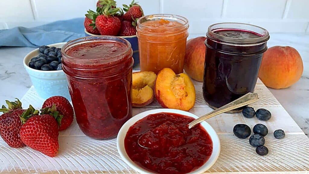 Three jars of fruit jam are displayed with fresh strawberries, blueberries, and peaches, alongside a bowl of jam with a spoon on a white board.