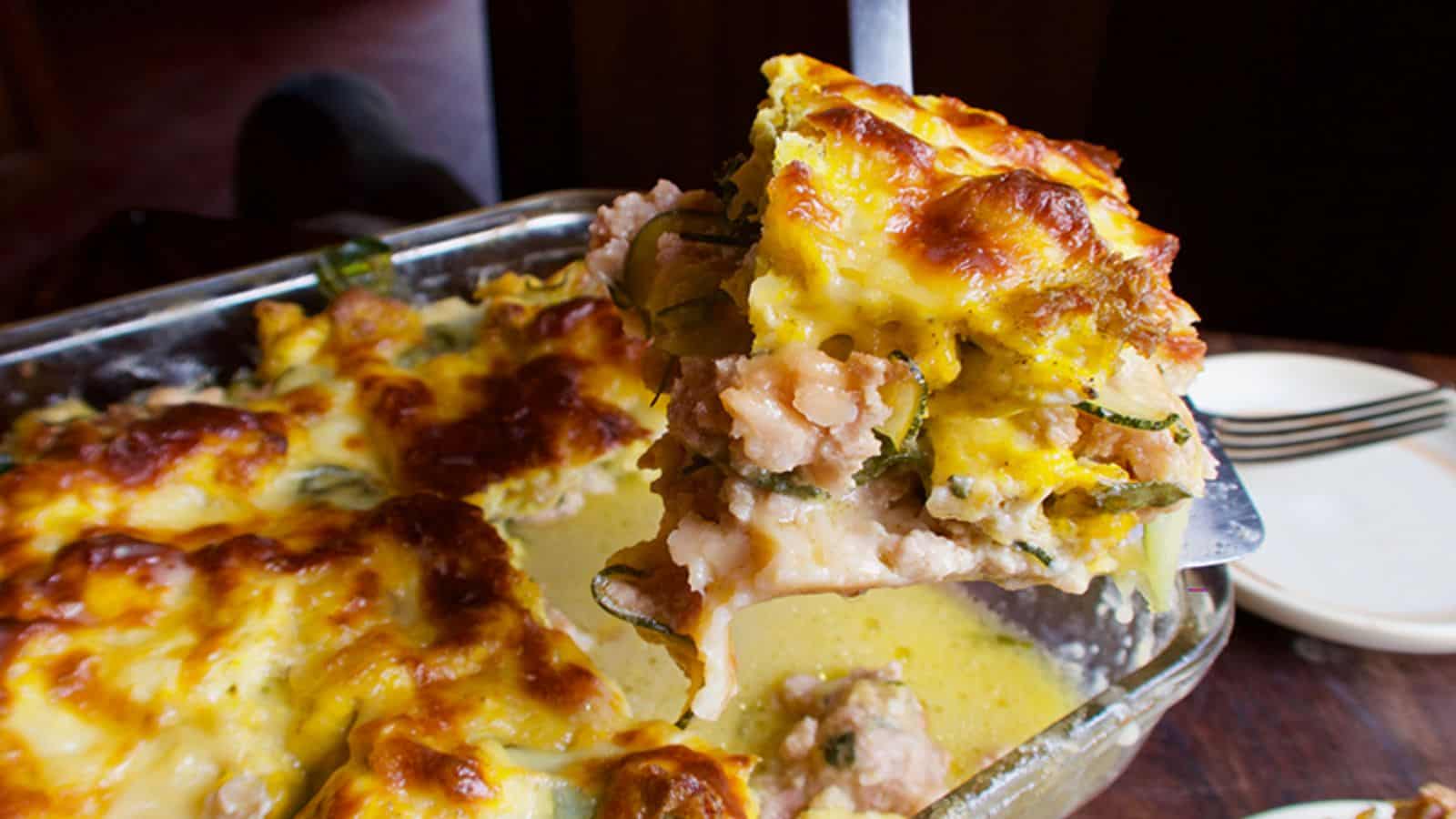 A serving of cheesy, baked casserole with vegetables and meat—perfect for high-protein breakfasts—is being lifted from a glass baking dish.