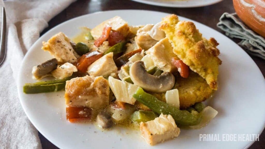 A white plate with baked chicken, mushrooms, green peppers, onions, tomatoes, and pieces of golden biscuit topping.
