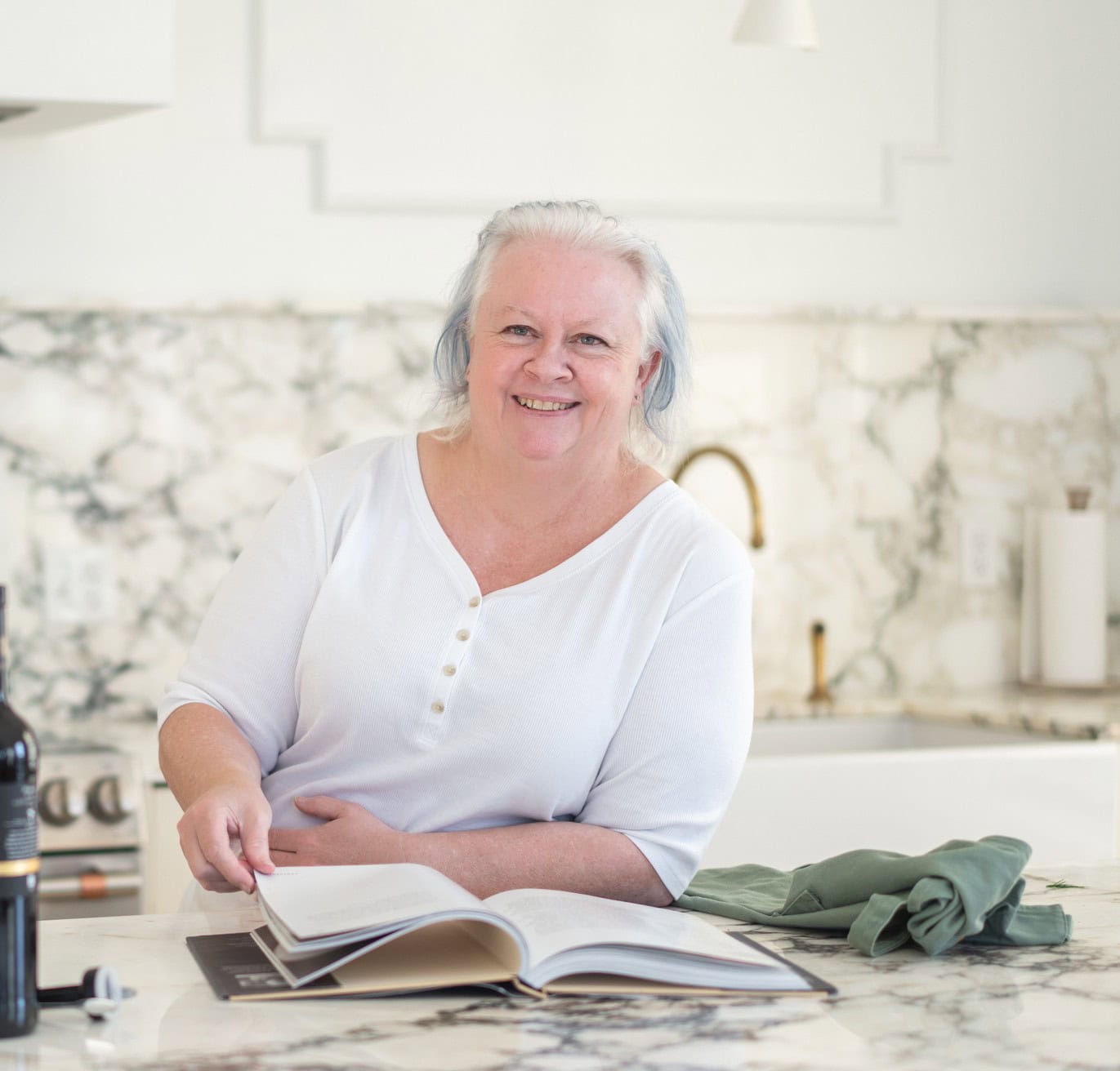 Older woman with gray hair sits at a marble kitchen counter, smiling while flipping through a cookbook. A wine bottle and folded green cloth are visible on the counter.