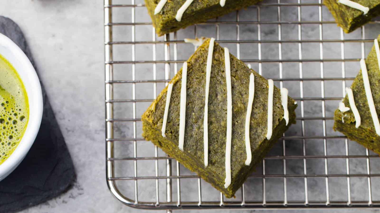 Square green scones with white icing drizzle cooling on a wire rack next to a cup of green beverage on a gray surface.